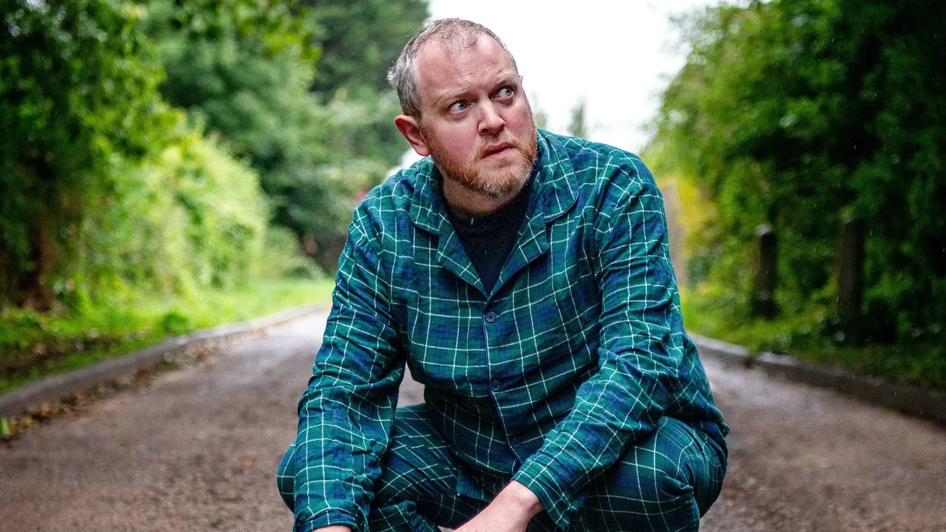 Miles Jupp smiling, wearing a blue shirt, against a blurred background with soft lighting.