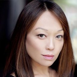 Naoko Mori performing on stage, wearing a black dress, with dramatic lighting and an attentive audience.