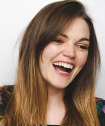 Naomi Cooper smiling, wearing a blue blouse, with long brown hair and a neutral background.
