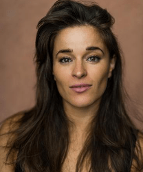 Natasha O'Brien smiling, wearing a blue blouse, with long brown hair and a neutral background.