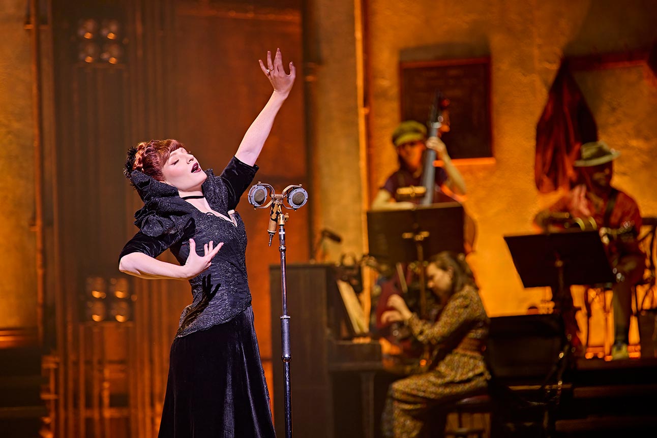 Nicola Roberts performs in a black velvet dress, illuminated by warm stage lighting, with musicians behind.