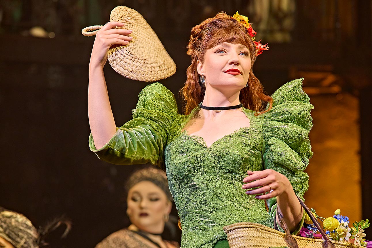 Nicola Roberts as Persephone, wearing a vibrant green dress, holding a straw hat and flowers.