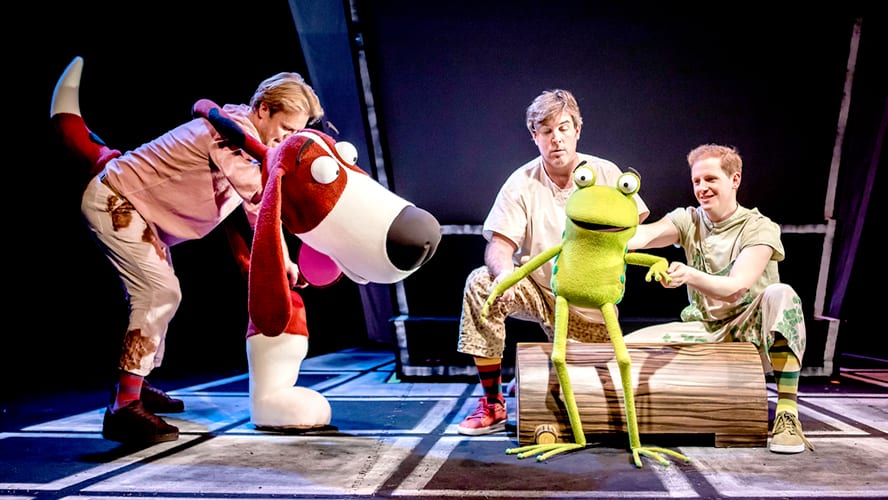 Colourful stage scene featuring animal puppets, including a frog, cat, and dog, in a playful setting.