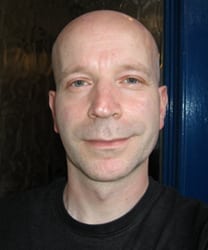 Oliver Latka smiling, wearing a dark suit and tie, with a blurred background.