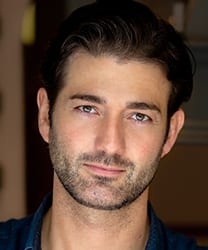 Oliver Tompsett smiling, wearing a black jacket, with short dark hair and a neutral background.