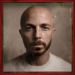 Bald man with a beard, wearing a white shirt, against a textured, sepia-toned background.