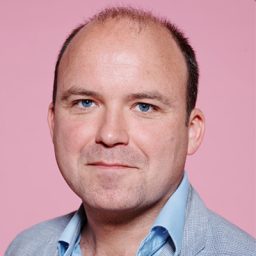 Rory Kinnear performing on stage, wearing a black shirt and standing in front of a spotlight.