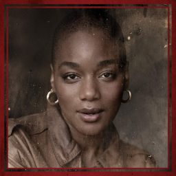 A woman with hoop earrings and a brown top, looking directly at the camera, neutral background.