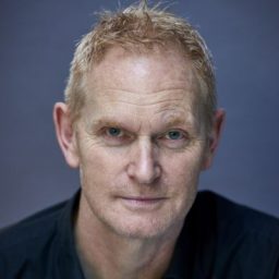Rob Alexander-Adams, a man with short dark hair, smiling against a blurred outdoor background.