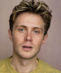 Rob Houchen smiling, wearing a dark suit and tie, with a blurred background.