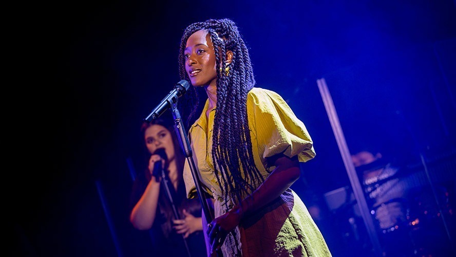 A diverse group of actors rehearsing on stage, with dramatic lighting and set pieces in the background.