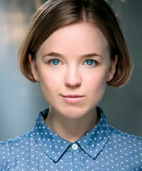 Rosie Abraham smiling, wearing a blue blouse, with shoulder-length brown hair and a neutral background.