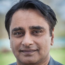 Sanjeev Bhaskar smiling, wearing a black shirt, with a blurred background.