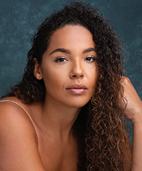 Shanay Holmes smiling, wearing a blue blouse, with long dark hair and a neutral background.