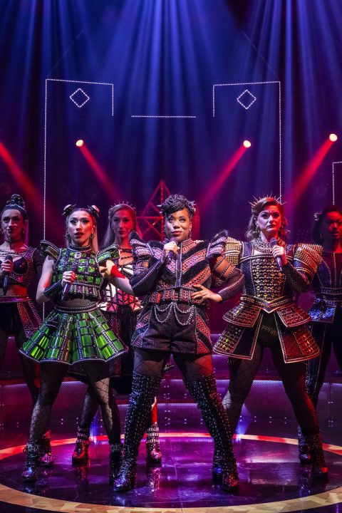 Cast members of SIX the Musical performing on stage, dressed in vibrant costumes with dramatic lighting.