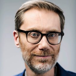 Stephen Merchant standing on stage, smiling, with a microphone in hand and spotlight above.