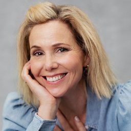 Sally Phillips smiling, with shoulder-length hair and a black top against a neutral background.