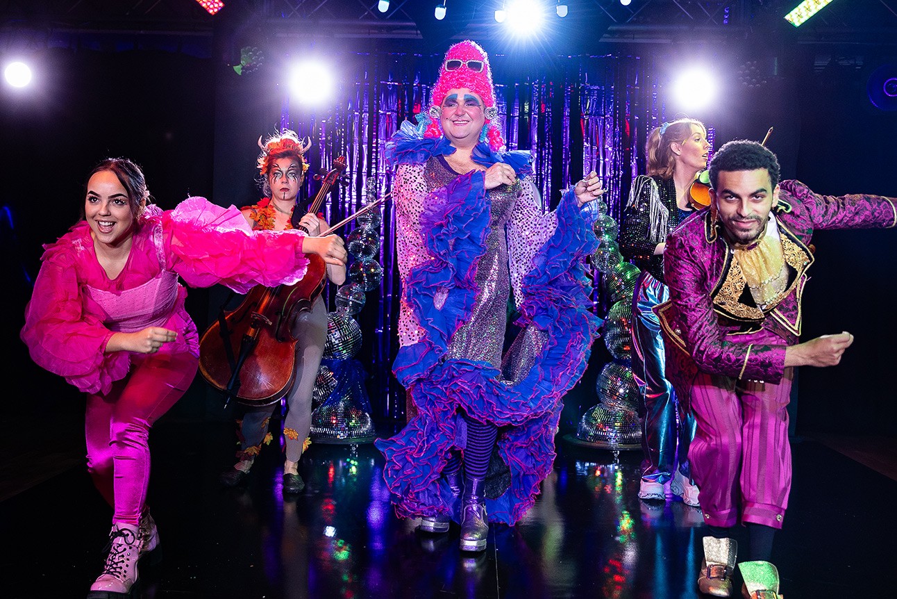A stage scene featuring actors in dramatic costumes, illuminated by soft lighting and smoke effects.
