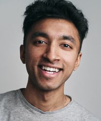 Shane David Joseph smiling, wearing a navy suit and white shirt, with a neutral background.
