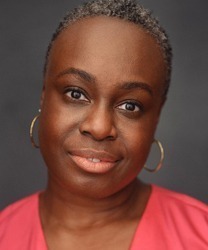 Sharlene Hector smiling, wearing a blue blouse, with long dark hair and a neutral background.