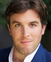 Simon Yadoo smiling, wearing a dark suit and tie, with a blurred background.