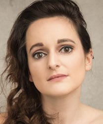 Sooz Kempner smiling, wearing a black top, with long brown hair and a neutral background.