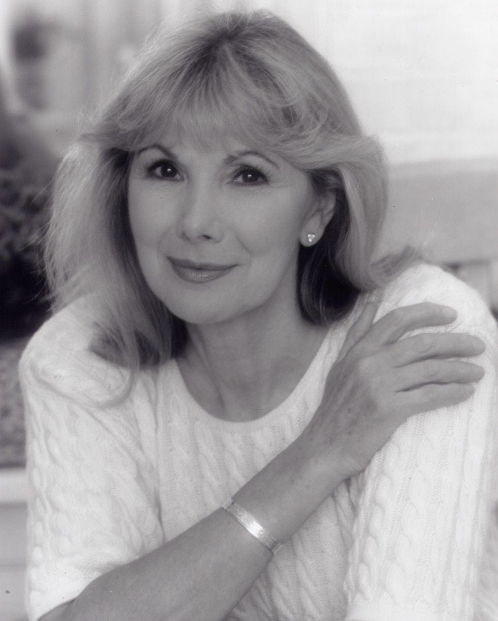 Susan Hampshire smiling, wearing a blue blouse, with shoulder-length brown hair and a neutral background.