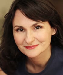 Suzy Bloom smiling, wearing a blue blouse, with shoulder-length brown hair and a neutral background.
