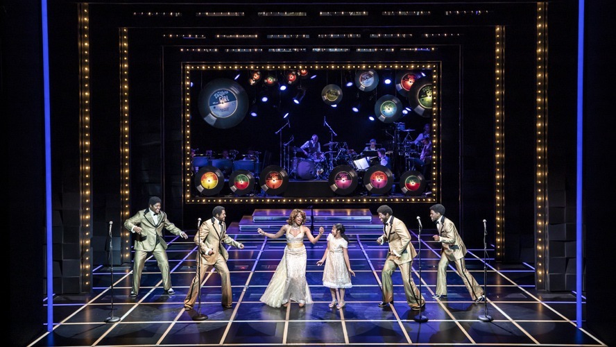 A vibrant stage scene featuring performers in colourful costumes, singing and dancing energetically.