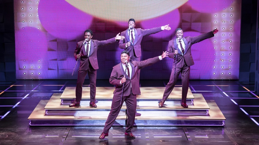 A vibrant stage scene featuring performers in 1960s attire, singing and dancing energetically.