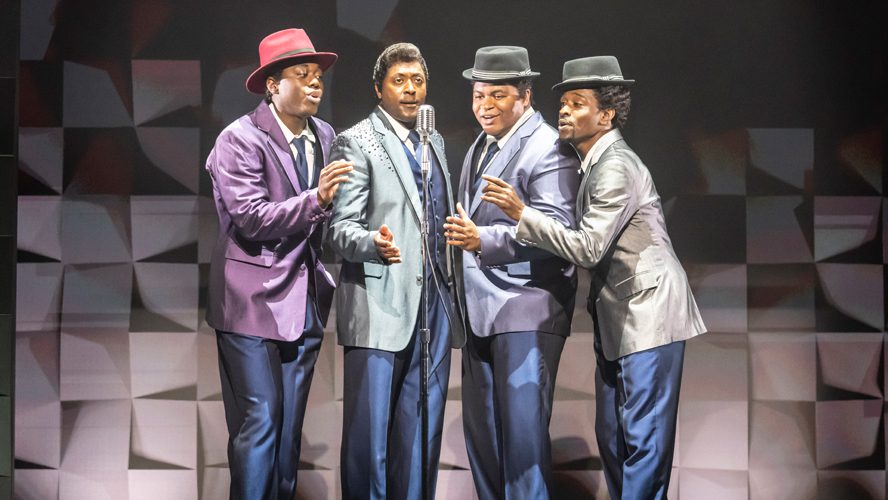 A vibrant stage scene featuring performers in colourful costumes, singing and dancing energetically.