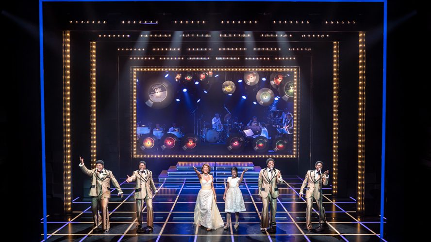 A vibrant stage scene featuring performers in colourful costumes, singing and dancing energetically.
