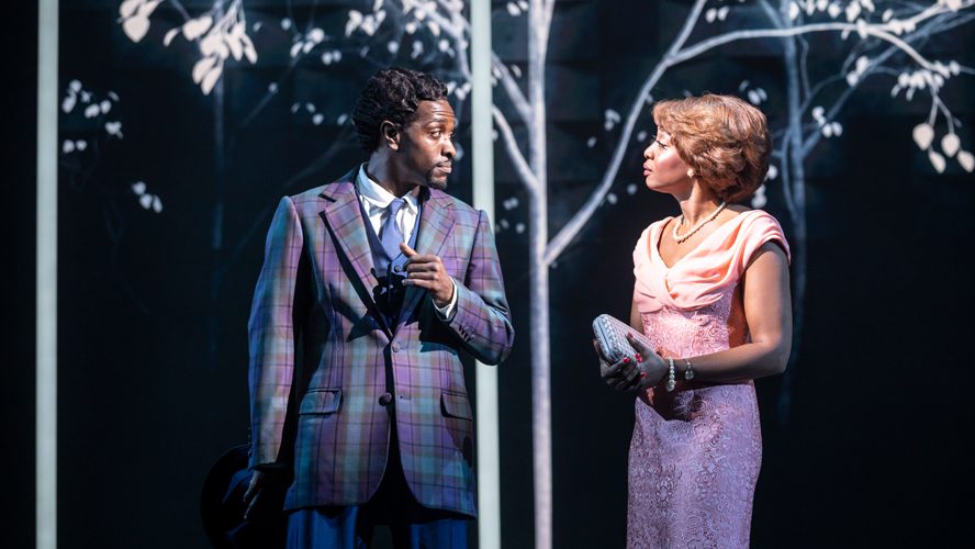 A vibrant stage scene featuring performers in 1960s attire, singing and dancing energetically.