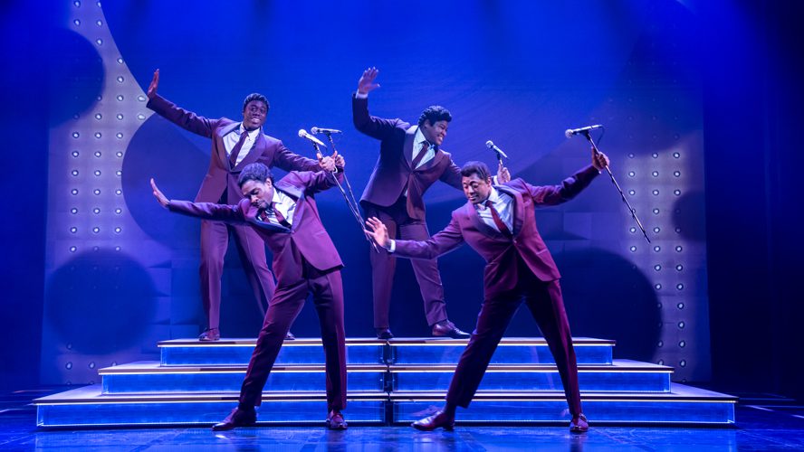 A vibrant stage scene featuring performers in stylish costumes, with dramatic lighting and a lively atmosphere.