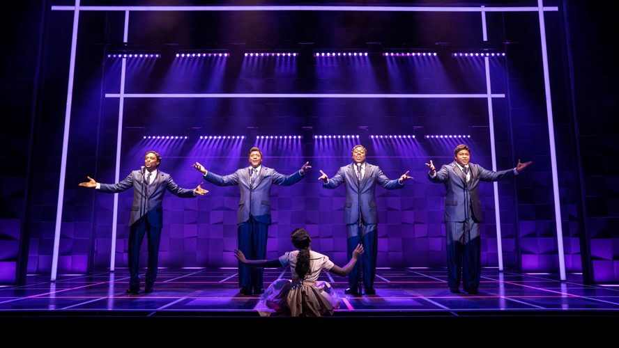 A vibrant stage scene featuring performers in colourful costumes, singing and dancing energetically.