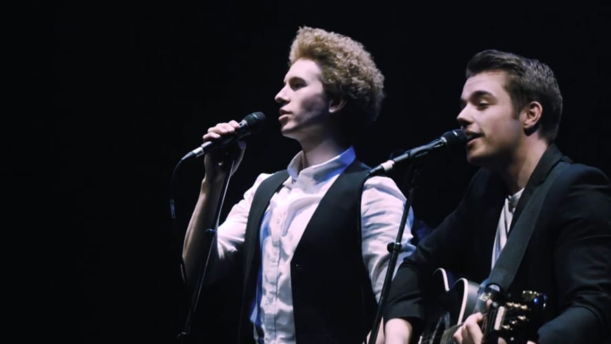 Simon and Garfunkel performing on stage, with microphones and a spotlight illuminating them.
