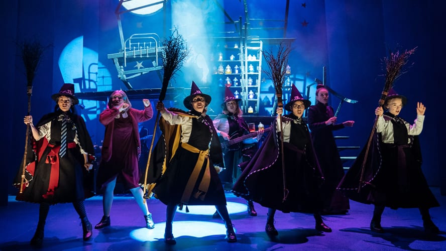 A young witch in a black robe stands in a classroom, surrounded by magical books and potions.