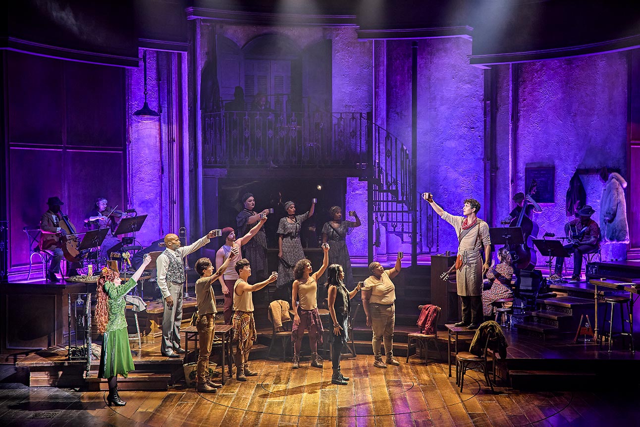 The cast of Hadestown performs on stage, illuminated by purple lights, with wooden flooring and instruments.