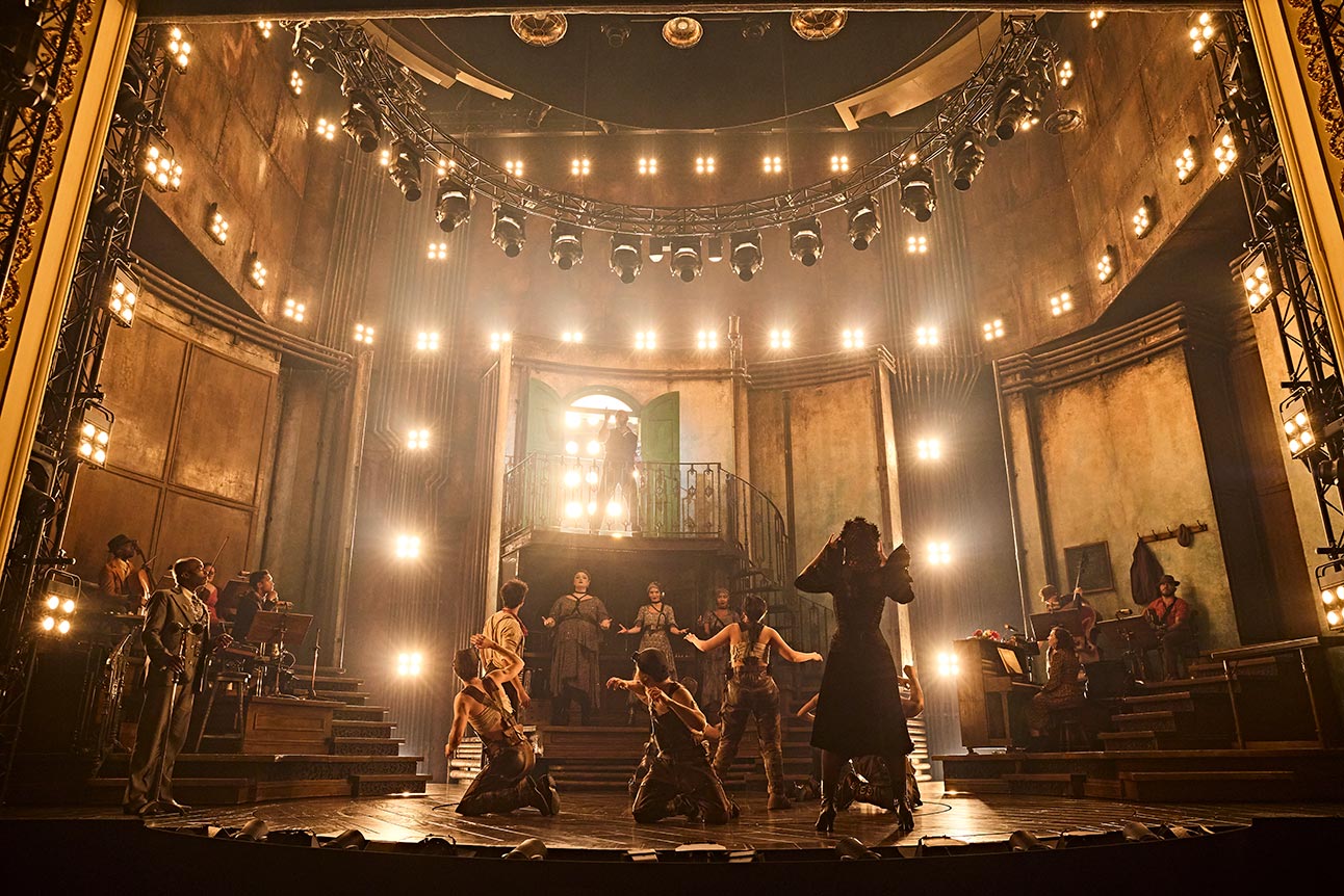 The cast of Hadestown poses together, wearing vibrant costumes, with a theatrical backdrop and dramatic lighting.