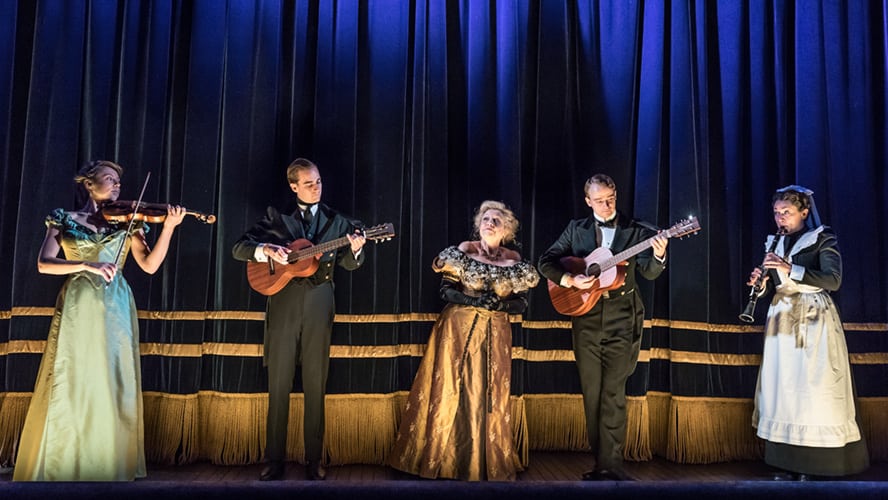 A woman playing a violin, surrounded by musicians, in a theatrical setting.