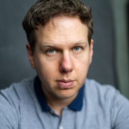 Thomas Coombes, a man with short dark hair, wearing a black shirt against a neutral background.