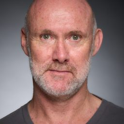 Close-up portrait of a man with a grey beard and short hair, wearing a dark grey shirt.