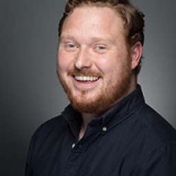 Tom Aldridge smiling, wearing a dark suit, with a blurred theatre backdrop behind him.