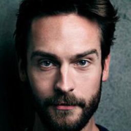 Tom Mison smiling, wearing a black jacket, with short dark hair and a light background.