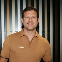 Dermot O'Leary smiling, wearing a dark suit and tie against a neutral background.