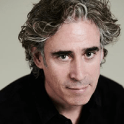 Stephen Mangan smiling, wearing a dark suit and tie, against a neutral background.