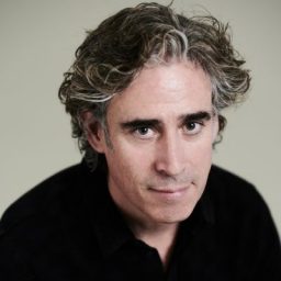 Stephen Mangan smiling, wearing a black shirt against a plain white background.