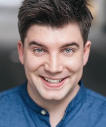 Charlie smiling, wearing a blue shirt, with short brown hair and a blurred outdoor background.