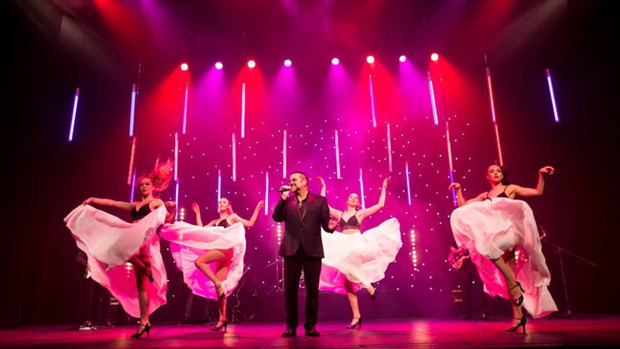 A vibrant stage with performers in stylish costumes, illuminated by colourful lights during a lively scene.