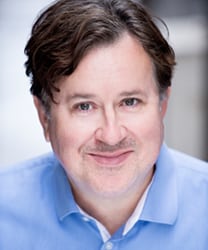 Jeremy Swift smiling, wearing a dark suit and tie, with a blurred background.